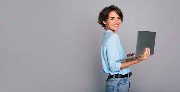 Banner Image of gorgeous young business woman in smart casual wear with cool hairstyle standing isolated over gray background and using her laptop computer and looking on camera.
