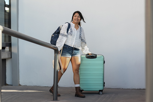 Horizontal photograph of a beauty young woman traveling alone through Mexico in her 20s, arriving at the bus station with her suitcase, walking pulling her luggage with a smile, excited to explore