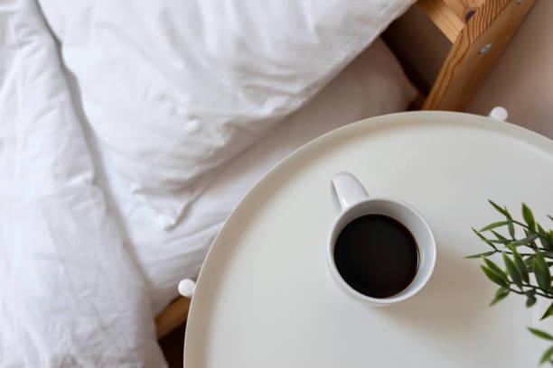 White bedside table with coffee cup and green plant, unmade bed with white cotton bedding. Simple bedroom interior design, morning coffee concept.