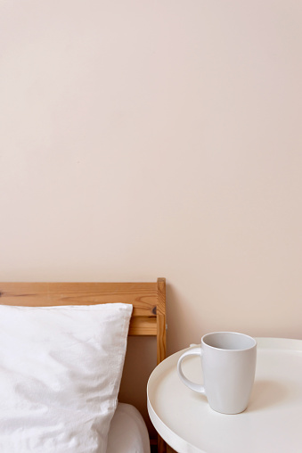 Minimalist simple lifestyle bedroom design interior. Bed with white linen pillow and blanket, bedside table with cup of coffee, empty neutral light peach wall background with copy space.