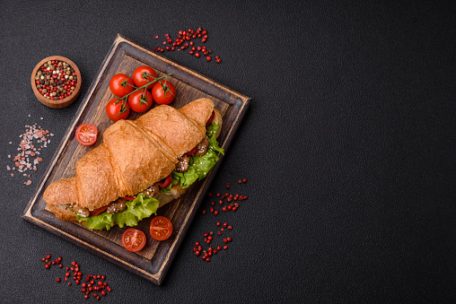 Delicious fresh crispy croissant with chicken or beef meat, lettuce, tomatoes, spices and sauce on a dark concrete background
