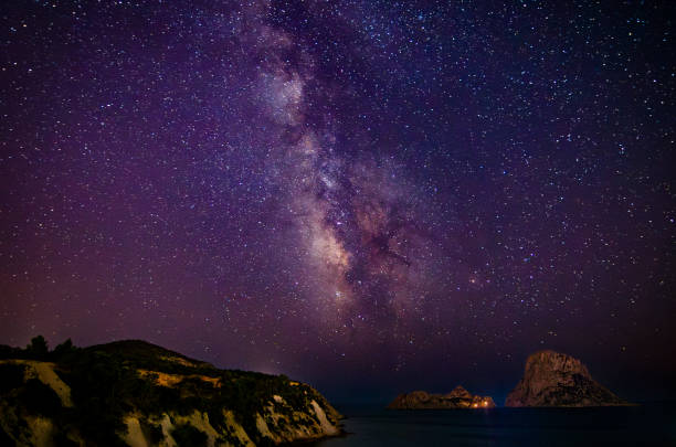 An image capturing the breathtaking Milky Way as it stretches across the night sky over the mystical island of Es Vedra, off the coast of Ibiza. This celestial scene highlights the natural beauty and cosmic wonder above this iconic landmark, known for its dramatic silhouette and spiritual allure.