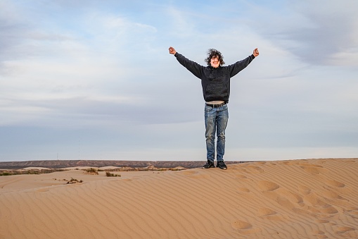 A male teenager raises his arms in joy and victory on top of a Sand Dune in the Chihuahua Desert at Lincoln National Forest, a national park covering 1.1 million acres and three mountain ranges in southeastern New Mexico administered by the US Forest Service.