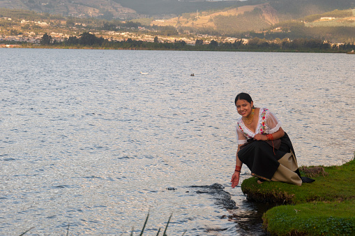 A young woman in traditional attire stands by the shore of a lake, her hands touching the surface as a warm glow softens the distant hills. Her expression embodies the joy of discovery as the day progresses toward the golden hour.