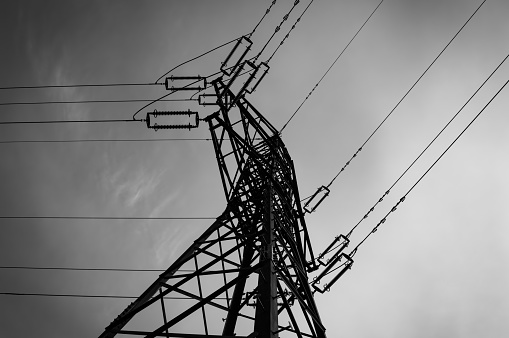 Monochrome image of a tangled network of power lines and a mast against a cloudy sky, highlighting industrial progress and energy distribution.