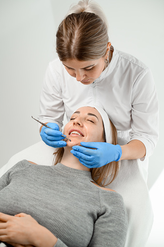 Beauty specialist in white overalls and blue rubber gloves makes a facial procedure with the help of cosmetic tools for a girl with a smile on her face lying on a couch