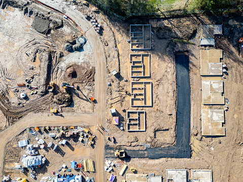 An aerial drone view looking down on new house foundations, framed out ready to be built up. The greenfield housing development is creating new homes in a rural location, expanding urban development. A dumper truck and stacks of construction material are located nearby, ready for builders to start the building of the modern homes.