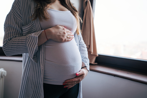 Young pregnant woman standing behind the window and holding hands on her belly.  Close up photo of female belly.