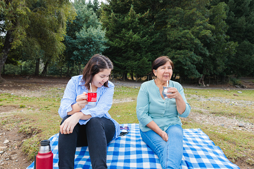 Generational Tradition: Latin Mother and Adult Daughter Share Yerba Mate on Blanket in Countryside Landscape