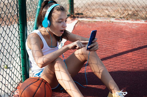 Portrait of a beautiful teenage girl reading surprising news on social media on her smartphone. A basketball - ball next to her.