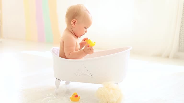 a little baby girl of six months with blue eyes bathes in a bubble bath and plays with rubber toys with ducks, smiles and rejoices splashing in the water, a happy baby washes