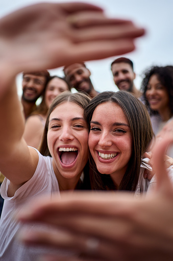 Vertical photo. Two girl friends making a frame with hands. A group of young people is happily celebrating, taking a selfie together. Smiling women having fun during their leisure travel