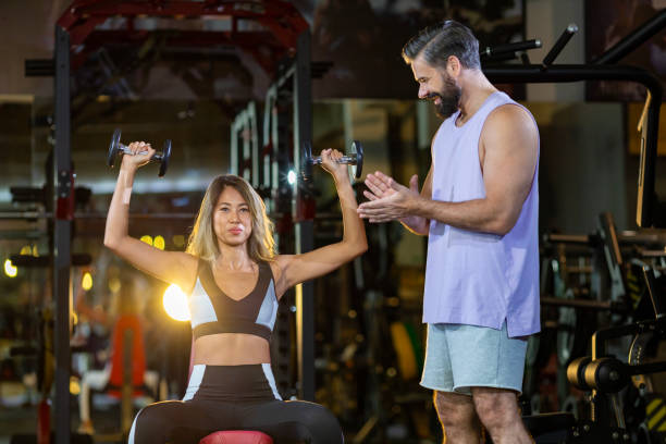 la donna asiatica sta usando il manubrio nell'allenamento con i pesi per il muscolo del braccio all'interno della palestra con supporto e applausi dall'allenatore con sfondo scuro per l'esercizio e l'allenamento - dumbbell weights weight training biceps femoris foto e immagini stock