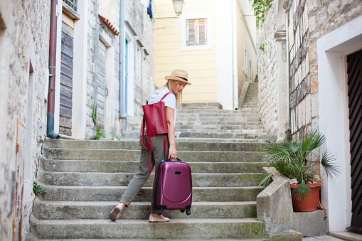 Girl traveler holds heavy suitcase on high stairs. Woman tourist carries luggage at old town street to authentic apartments. Concept of travel, summer vacation, solo female tourism, trip, adventure.