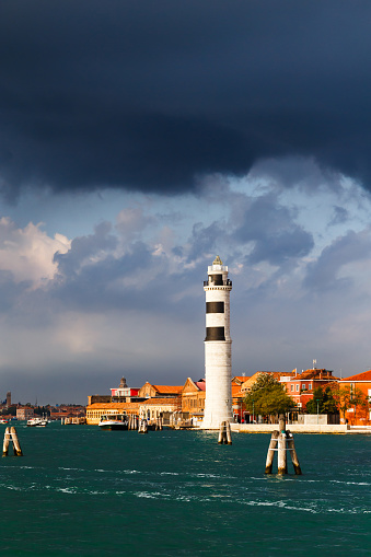 Murano Lighthouse or Faro di Murano is active lighthouse located on Murano island, Venice, Italy, in Venetian Lagoon on Adriatic Sea. View from water bus with dark blue stormy sky in background.