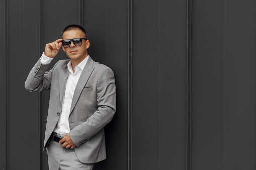 Handsome successful young man wearing gray suit, white shirt, sunglasses and posing against gray wall in modern city.