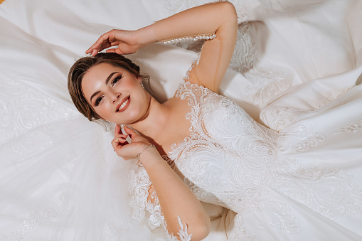 Beautiful bride, attractive woman in wedding dress, lying on bed. Fashionable hairstyle and make-up. Happy bride woman. Smiling bride. Wedding day. A gorgeous bride. Marriage.