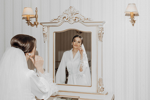 Portrait of a young beautiful bride in a beautiful hotel interior preparing for her wedding. A gorgeous woman looks in the mirror at her makeup and hairdo