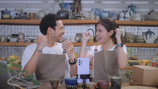 SME owners express joy over successful product orders during a live stream in their ceramic shop filled with handmade pieces. Southeast Asian and Thai ethnicity model, Males and Females, including white people, 25-29 years