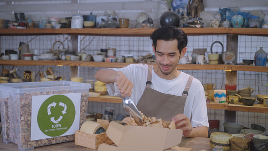 SME business owner chooses to use eco-friendly packaging, utilizing recyclable cardboard and water hyacinth cushioning for their pottery, showcasing an environmentally conscious and waste reduction approach. Southeast Asian and Thai ethnicity model, Males, 25-29 years