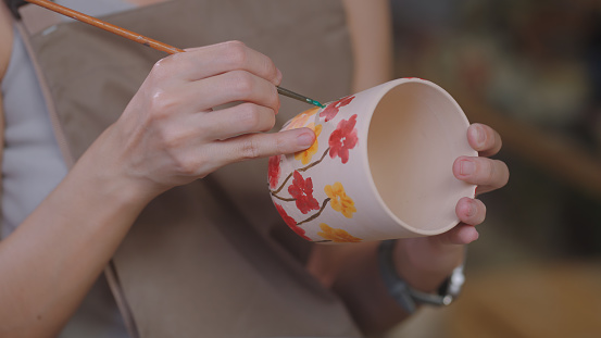 Close-up view of an artisan's hands delicately painting on a ceramic mug, adding artistic details to their ceramic creations in a well-stocked craft studio