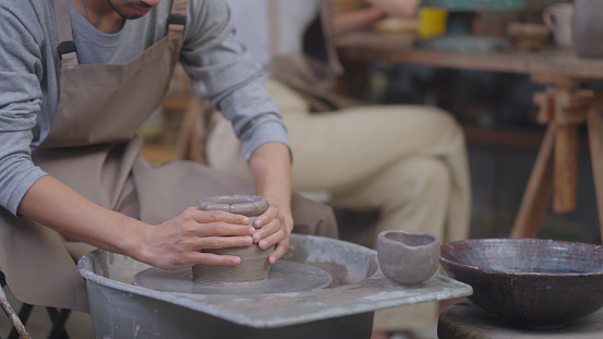 Hands of a potter masterfully shape clay on a spinning potter's wheel, creating a new piece in a pottery studio filled with earthenware