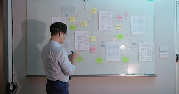 rear view asian senior businessman is putting his ideas and memorandum on whiteboard while overtime working in office