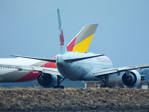 Asiana and Air Canada planes queued up for takeoff at Sydney Airport.  On the left is an Asiana Airlines A350-941 plane, registration HL8521, preparing to depart as flight OZ602 to Seoul.  On the right is an Air Canada Boeing B777-233(LR) plane, registration C-FIVK, preparing to depart as flight AC34 to Vancouver.  In the foreground are rocks forming part of the sea wall around the runway which juts out into Botany Bay.  This image was taken from near Kyeemagh Beach, Botany Bay on an overcast and rainy morning on 17 March 2024.