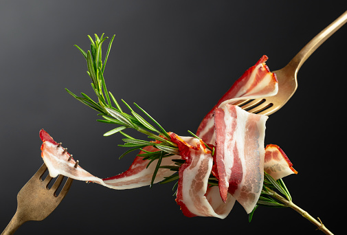 Dry-cured pork belly bacon with rosemary on a black background. Sliced bacon on a forks.