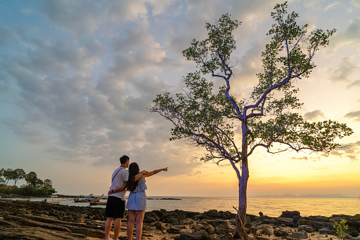 A couple standing on a beach looking at a tree. The sky is cloudy and the sun is setting. Krabi, Thailand