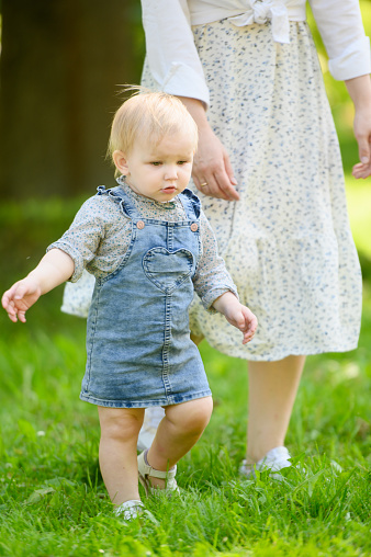 Toddler walks on the grass in the park next to his mother, learning to walk and take his first steps in nature. Development concept, first independent steps. Vertical.