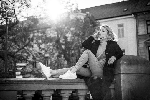 Back View of Woman in Black Coat and White Shoes Sitting on Ledge. Stylish Urban Fashion Portrait. Trendy Female Model Enjoying Cityscape. Casual Outdoor Lifestyle Concept.