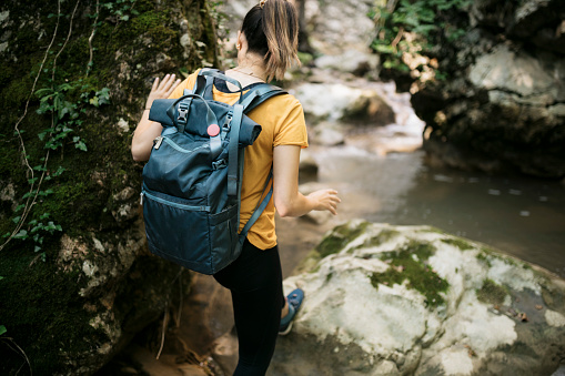 Rear view of a young woman on a hiking trip walking over big rocks along mountain creek. Female hiker with backpack hiking in forest.