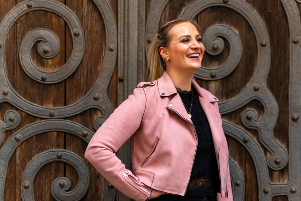 woman wearing a pink jacket and black pants stands in front of a wooden door stock photo