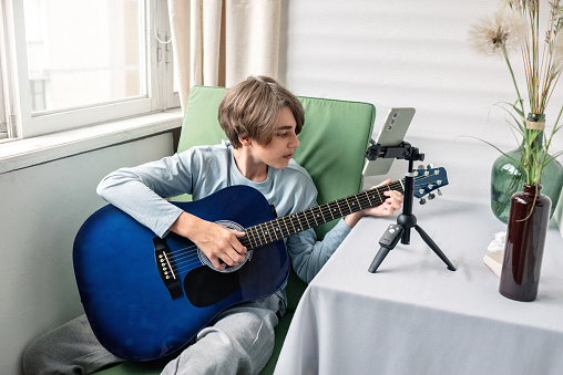 Teenager boy blogger playing guitar and recording video on smartphone, streaming online for social media blog or webinar lesson.
