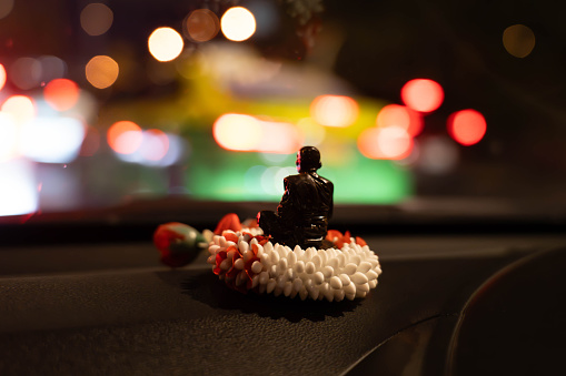Rear view of the small Buddha statue on the jasmine garland on the front of the car. Bokeh light background at night.