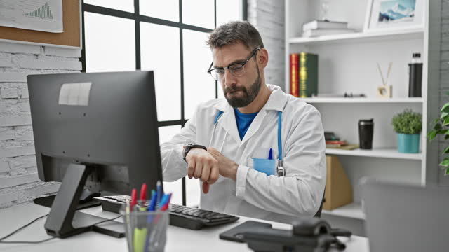 Handsome, bearded young hispanic man, a serious medical professional in clinic uniform, working indoors at a healthcare center. using smartphone, he looks at the clock, bothered by the ticking time.