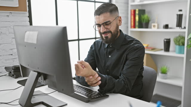 Bearded man with glasses in pain at office desk, clutching wrist in ergonomic discomfort with computer monitor in background.