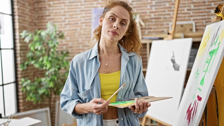 Confident woman artist holding palette and brush in a creative studio with easels and paintings