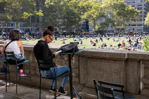 Manhattan, NY, USA - September 21, 2023: A young business woman with her hair in a bun and wearing jeans and sneakers sits on a chair at a park and looks at her phone. Her bag is on the table.