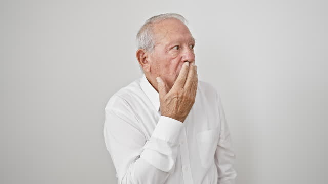 Shocked senior man with grey hair, standing in silence, hand covering mouth in fear and surprise. isolated white background portrays the intense emotion of this startled elder male.