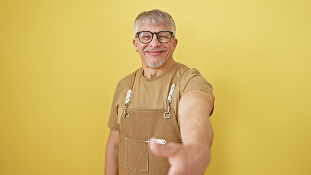 Smiling middle aged grey-haired man in glasses and apron extends hand for welcoming handshake. displays professional success over a yellow isolated background.