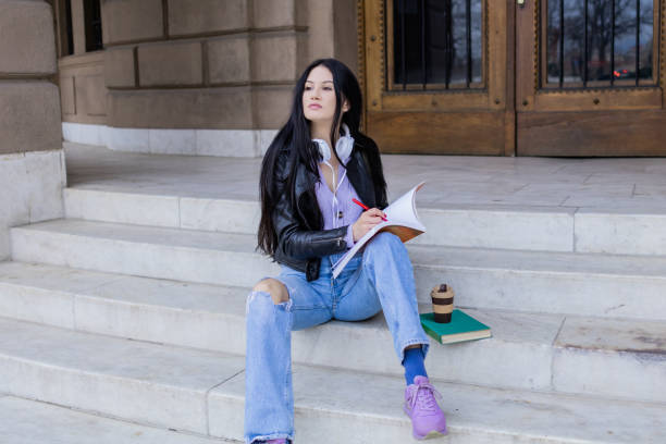 istock Young beautiful female university student with backpack sitting on college staircase and making notes with pencil, . She is wearing headphones and leather jacket while waiting for her college class to start. 2091120952