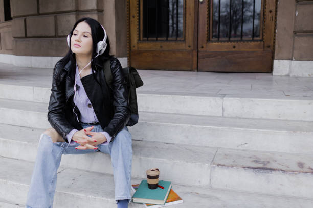 istock Young beautiful female university student with backpack sitting on college staircase. She is wearing headphones and leather jacket while waiting for her college class to start. 2091124609