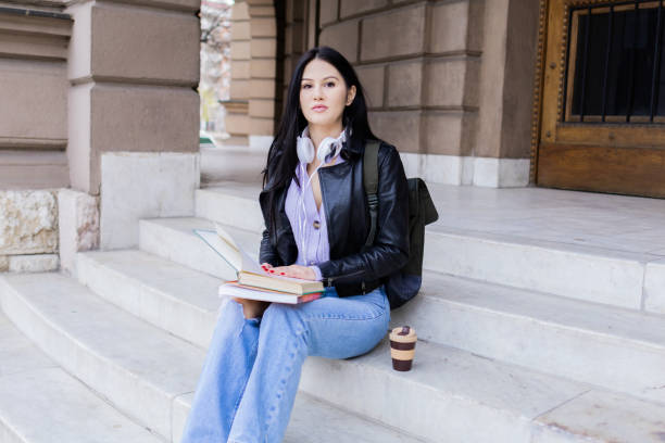 istock Young beautiful female university student with backpack sitting on college staircase. She is wearing headphones and leather jacket while waiting for her college class to start. 2091126205