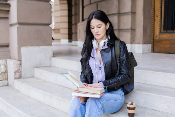 istock Young beautiful female university student with backpack sitting on college staircase. She is wearing headphones and leather jacket while waiting for her college class to start. 2091128258