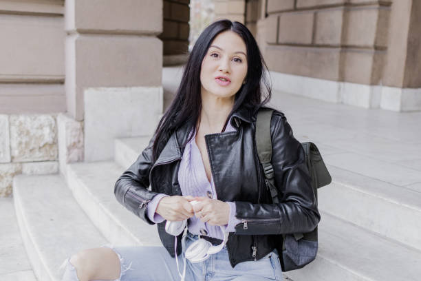 Young beautiful female university student with backpack sitting on college staircase. She is wearing headphones and leather jacket while waiting for her college class to start.