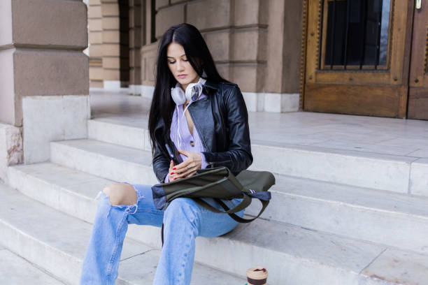 istock Young beautiful female university student with backpack sitting on college staircase. She is wearing headphones and leather jacket while waiting for her college class to start. 2091134006