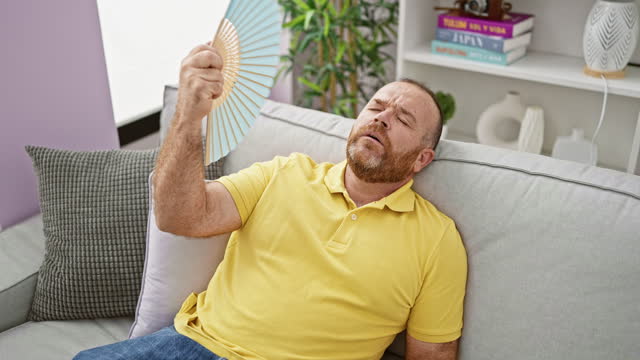 Middle-aged caucasian man suffering in hot weather, fanning himself with handfan while sitting on sofa at home