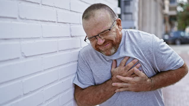 Handsome middle age caucasian man suffering heart attack outdoors on a sunny city street, emotion of pain evident as he clutches chest, leaning against wall with a look of sickened love.